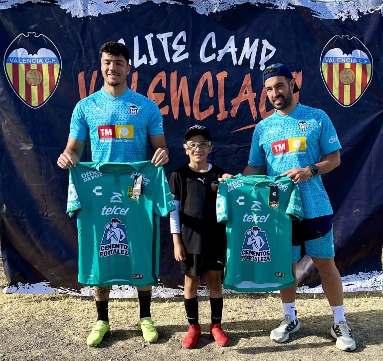 Dos entrenadores con uniformes azules del Valencia CF posan junto a un niño con uniforme negro. Los entrenadores sostienen camisetas verdes de fútbol. Detrás de ellos hay una pancarta que dice "ELITE CAMP VALENCIA CF" con el logo del club. Están sobre césped seco.
