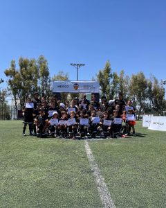 Grupo de niños y jóvenes en un campo de fútbol, sosteniendo certificados, con un banner de Valencia CF Soccer Camps al fondo.