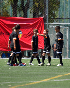 Grupo de niños en un campo de fútbol, vestidos con camisetas negras y detalles naranjas, interactuando y sonriendo, con un fondo de una pancarta roja que dice "México".