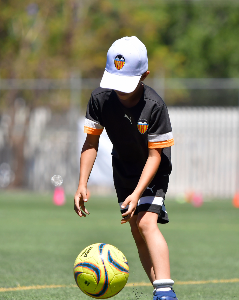 Niño en un campo de fútbol, vestido con camiseta negra y gorra blanca, agachándose para patear un balón de fútbol amarillo con detalles de colores.