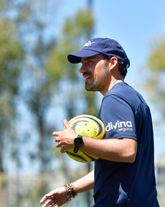 entrenador en un campo de fútbol, vestido con camiseta y gorra azul marino, sosteniendo balón de fútbol amarillo con detalles de colores.