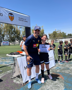 Entrenador y niña en un campo de fútbol, sosteniendo certificados, con un banner de Valencia CF Soccer Camps al fondo.