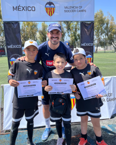 Entrenador y grupo de niños en un campo de fútbol, sosteniendo certificados, con un banner de Valencia CF Soccer Camps al fondo.