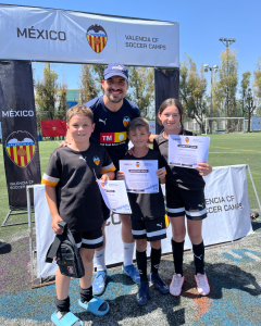 Entrenador y grupo de niños en un campo de fútbol, sosteniendo certificados, con un banner de Valencia CF Soccer Camps al fondo.